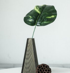 Tall four-sided tapered vase with textured grey panels holding a single leaf, beside a dried lotus pod