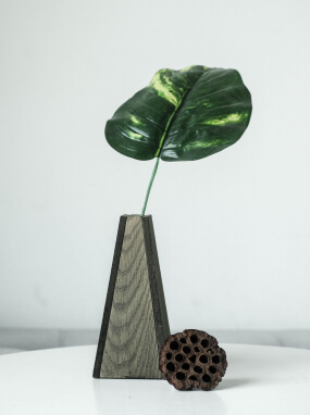 Tall four-sided tapered vase with textured grey panels holding a single leaf, beside a dried lotus pod